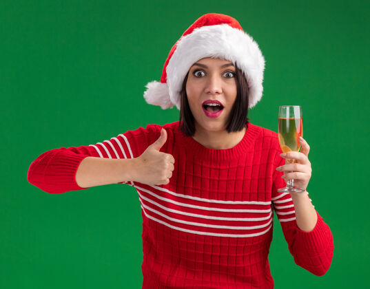 展示令人印象深刻的年轻女孩戴着圣诞帽拿着一杯香槟在绿色的墙上孤立地竖起大拇指圣诞节拇指香槟