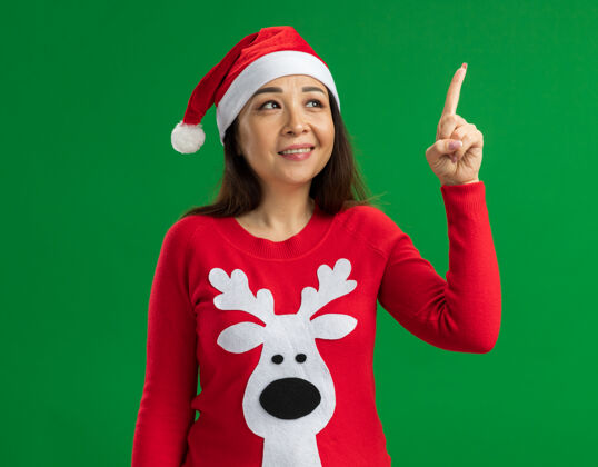 手指快乐的年轻女子戴着圣诞老人帽 穿着红色毛衣 站在绿色的背景上 用食指指着头 有了新的想法指向有索引