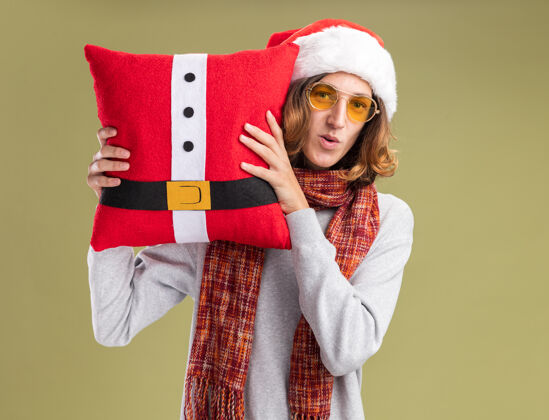 围着年轻人戴着圣诞老人帽 戴着黄色眼镜 脖子上围着暖和的围巾 抱着圣诞枕头 站在绿色的背景下 面带微笑地看着相机圣诞老人眼镜年轻