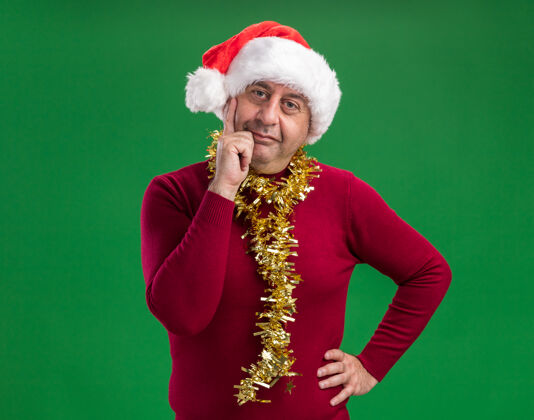 金属片中年男子戴着圣诞老人的帽子 脖子上戴着金属丝 站在绿色的背景下看着相机 自信地微笑着中间周围站立
