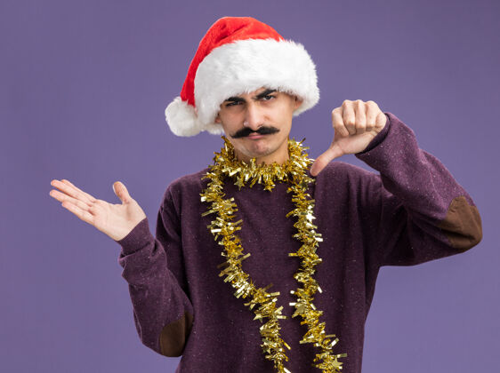 帽子年轻的胡子男人戴着圣诞老人帽子 脖子上戴着金属丝 不高兴地看着相机 大拇指朝下 胳膊站在紫色的背景上手臂脖子向下