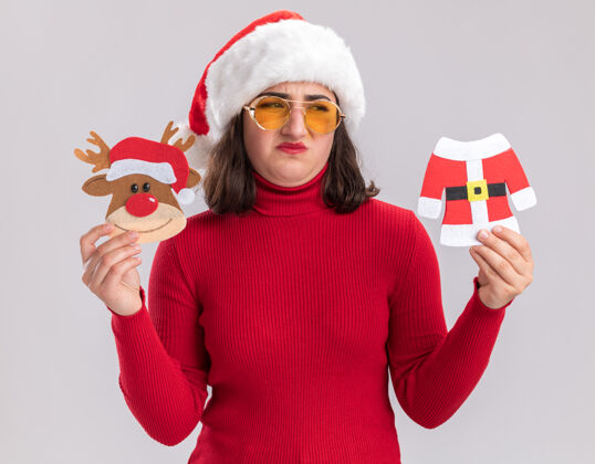 疑惑穿着红毛衣戴着圣诞帽戴着眼镜拿着圣诞玩具的小女孩站在白色的墙上困惑地看着一边有疑惑帽子圣诞节毛衣