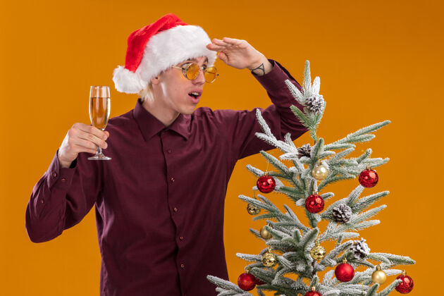 印象深刻令人印象深刻的年轻金发男子戴着圣诞帽和眼镜站在装饰圣诞树附近拿着香槟酒杯望着远方的橙色墙上帽子金发玻璃杯