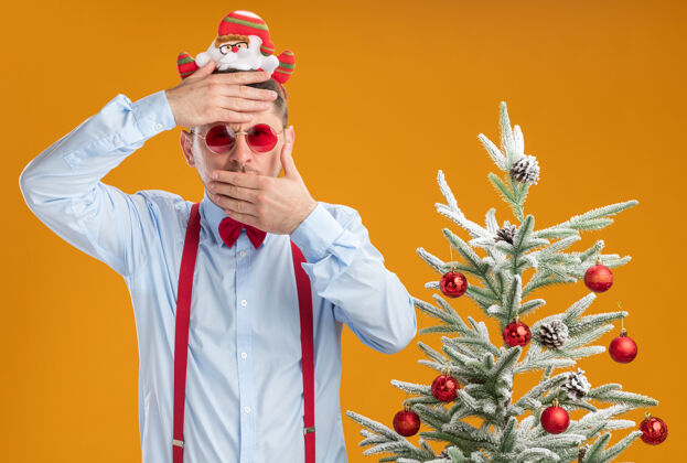 圣诞老人戴着吊带领结的年轻人站在圣诞树旁 戴着圣诞老人和红眼镜 一只手放在额头上 另一只手放在橘色的墙上 捂住嘴圣诞节轮辋穿
