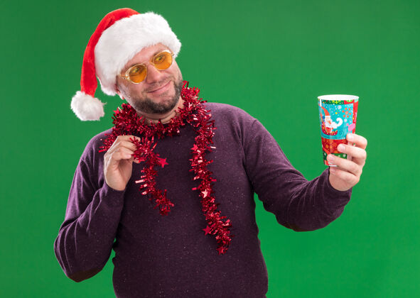 圣诞节高兴的中年男子戴着圣诞帽 脖子上戴着金属丝花环 戴着眼镜 伸出塑料圣诞杯 看着它触摸着隔离在绿色墙上的金属丝花环帽子高兴金属丝