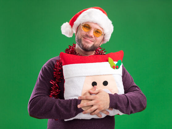 圣诞老人高兴的中年男子戴着圣诞帽 脖子上戴着金箔花环 戴着眼镜 把圣诞老人的枕头孤立在绿色的墙上金属丝周围高兴