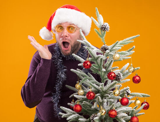 圣诞老人令人印象深刻的中年男子戴着圣诞帽 脖子上戴着金箔花环 戴着眼镜 站在装饰过的圣诞树后面 一只空手孤立在橙色的墙上男人站着周围