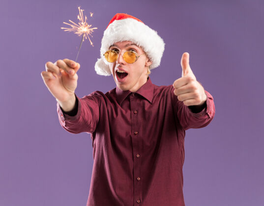 金发令人印象深刻的年轻金发男子戴圣诞帽和眼镜举行假日火花显示拇指向上孤立的紫色墙上帽子印象深刻圣诞老人