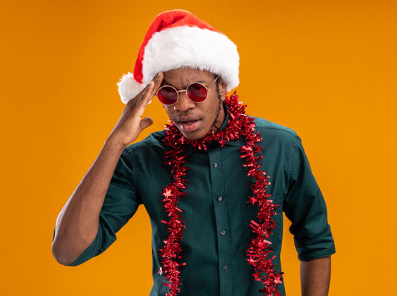头戴着圣诞帽 戴着花环 戴着太阳镜的非裔美国人站在橙色的墙上 困惑地看着一边 手放在头上圣诞老人非洲人花环