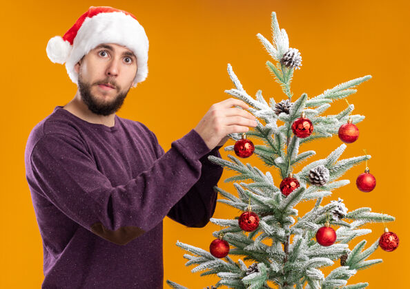 帽子一个穿着紫色毛衣 戴着圣诞帽的年轻人站在橙色的墙上 把玩具挂在圣诞树上 看起来很困惑男人困惑圣诞节
