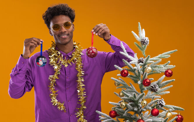 圣诞饰品微笑着的年轻的非裔美国人戴着眼镜 脖子上戴着金箔花环 站在橙色背景的装饰过的圣诞树旁附近金箔圣诞树