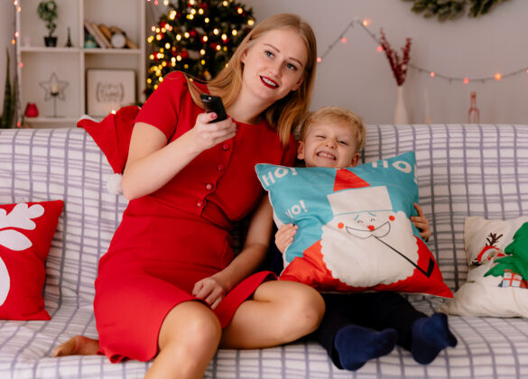 沙发快乐的妈妈穿着红色的衣服 她的孩子坐在沙发上 在装饰好的房间里一起看电视 背景是圣诞树房间电视圣诞