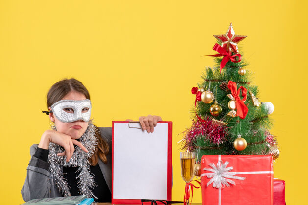 桌子前视图体贴的年轻女孩戴着面具坐在桌旁圣诞树和鸡尾酒礼物时尚体贴头发