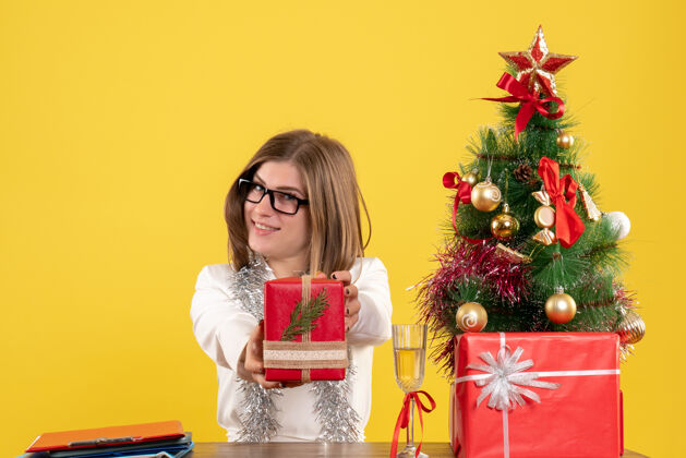桌子前视图：女医生坐在桌子前 拿着礼物 黄色背景上的圣诞树和礼品盒背景礼物快乐