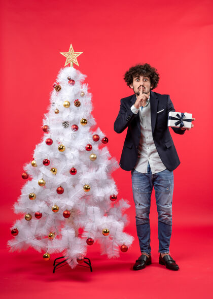 站着严肃的年轻人拿着他的礼物制作沉默的手势站在装饰过的白色圣诞树旁沉默手势年轻人