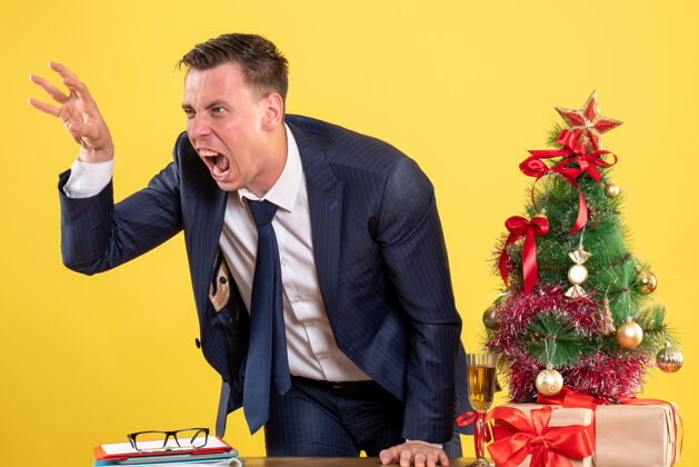 商人愤怒的男人站在圣诞树旁的桌子后面 在黄色的墙上展示礼物男性西装公司