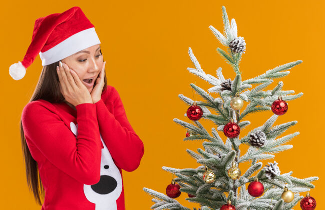 圣诞节惊讶的年轻亚洲女孩戴着圣诞帽 穿着毛衣站在圣诞树旁 两手被隔离在橙色的背景上附近戴着盖着