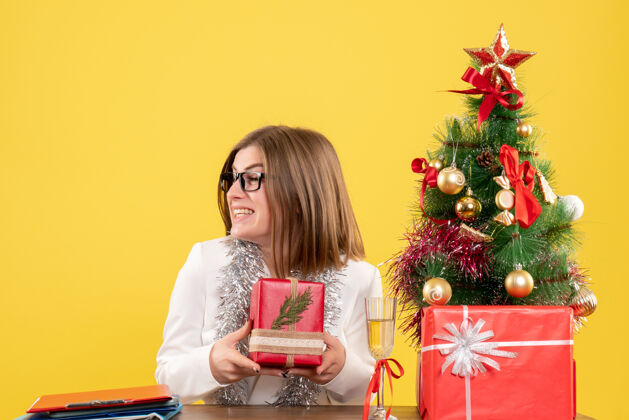 办公室正面图：女医生坐在桌子前 拿着礼物 黄色背景上的圣诞树和礼盒人礼物女性