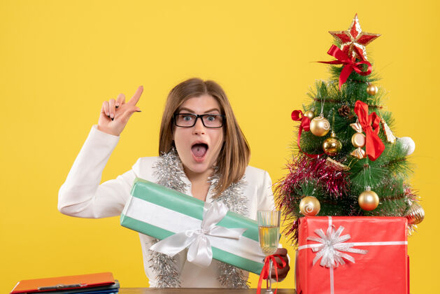 成人正面图：女医生坐在桌子前 拿着礼物 黄色背景上的圣诞树和礼盒树前面人