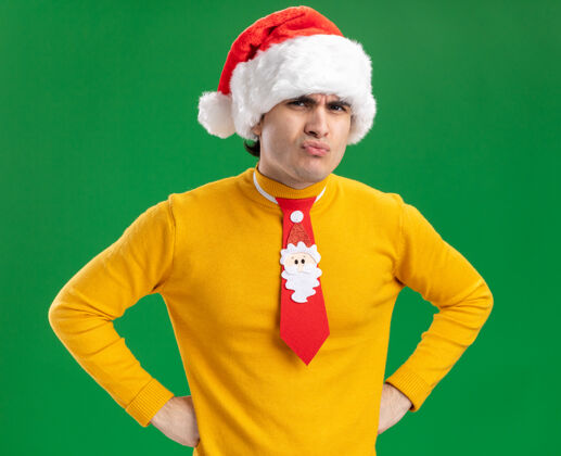 高领毛衣一个穿着黄色高领毛衣 戴着圣诞帽 打着滑稽领带的年轻人看着摄像机 不高兴地看着愤怒的脸 双手放在臀部 站在绿色的背景上搞笑圣诞老人男人