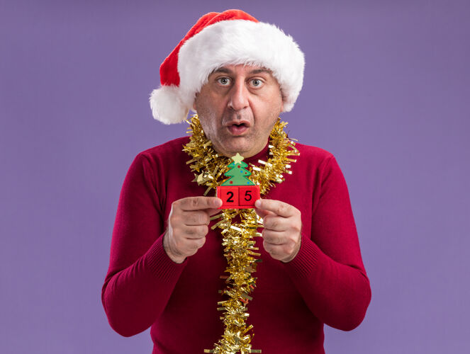 举行中年男子戴着圣诞老人帽子 脖子上戴着金属丝 手里拿着玩具方块和25岁的约会对象 站在紫色的背景下 看着相机 心神不安 不知所措日期二十立方体