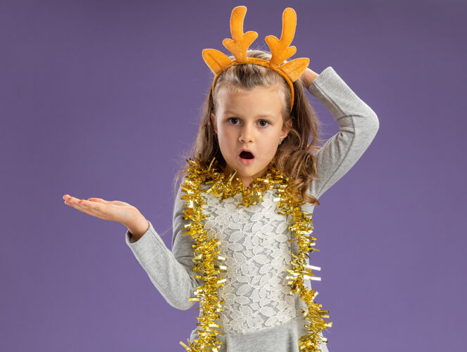 头发困惑的小女孩戴着圣诞发箍 脖子上戴着花环 手放在一边 手放在脑后 孤立在蓝色背景上箍快乐头