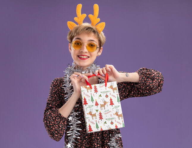 鹿角微笑的年轻漂亮女孩戴着驯鹿鹿角头带 脖子上戴着金箔花环 戴着眼镜 手里拿着圣诞礼品袋 看着紫色背景上孤立的相机相机礼物包