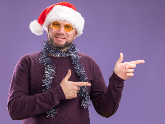 高兴一个戴着圣诞老人帽 脖子上戴着金箔花环 戴着眼镜的中年男人 站在紫色的墙上 脸上露出一副孤零零的眼镜穿着圣诞节周围
