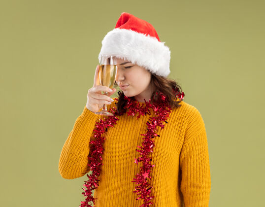 圣诞老人戴着圣诞帽 脖子上戴着花环 拿着一杯香槟酒 橄榄绿背景上有复制空间的未出租的年轻斯拉夫女孩年轻周围帽子
