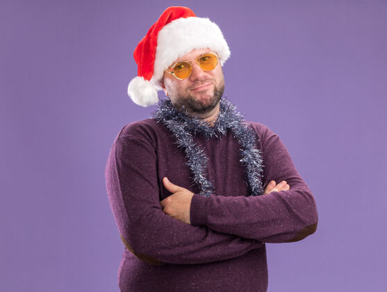 关闭高兴的中年男子戴着圣诞老人帽 脖子上戴着金箔花环 戴着眼镜 站在紫色的墙上 有复制空间周围帽子高兴