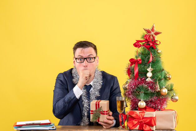 黄色前视图震惊的人拿着礼物坐在圣诞树附近的桌子上 黄色的礼物坐着男人人