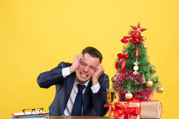 商人愤怒的男人抱着头坐在圣诞树旁的桌子前 黄色的墙上挂着礼物圣诞节树生气