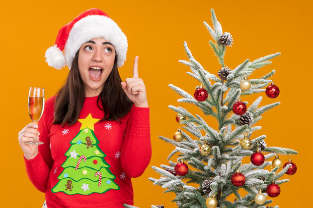 圣诞树一个戴着圣诞帽的年轻白人女孩拿着一杯香槟 惊讶地指了指帽子橙色香槟