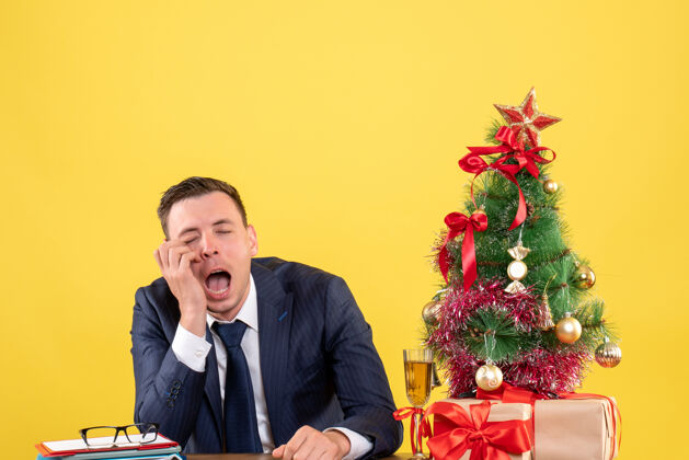 人抑郁的男人坐在圣诞树旁的桌子前 黄色的礼物男人西装坐着