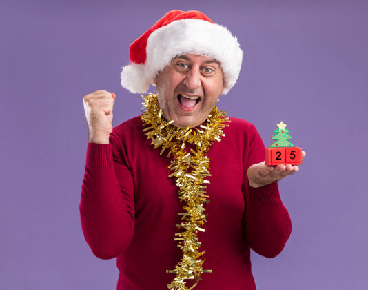 握紧中年男子戴着圣诞老人的帽子 脖子上戴着金属丝 手里拿着玩具方块和日期 二十五岁快乐而兴奋地握紧拳头站在紫色的背景上立方体周围拳头