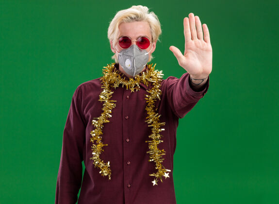 圣诞节严肃的年轻金发男子戴着眼镜 戴着防护面具 脖子上戴着金属丝花环 在绿色的墙上做着停止手势穿严重做