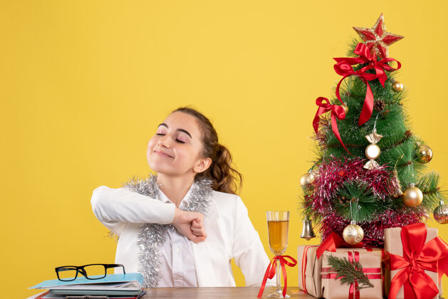 微笑前视图：女医生坐在桌子后面 拿着圣诞礼物和黄色背景上的树圣诞节插花医生