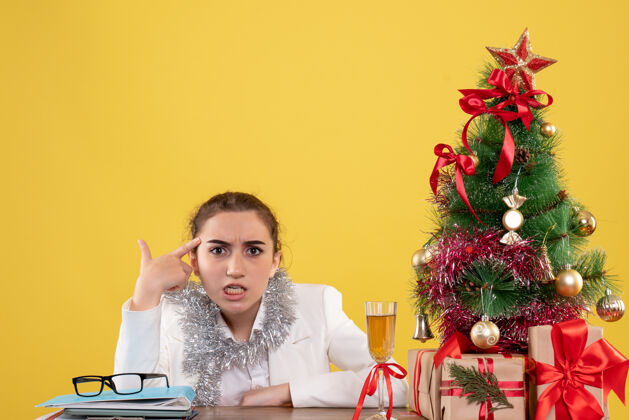情感正面图：女医生坐在黄色背景的桌子后面 还有圣诞树和礼品盒坐着女性花