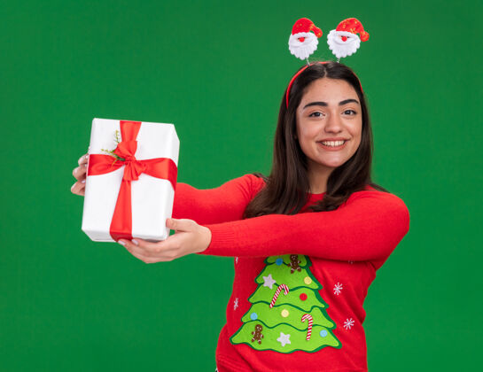 盒子微笑着的年轻白人女孩 戴着圣诞老人的头带 手拿着圣诞礼盒 绿色背景上有复制空间头带微笑礼物