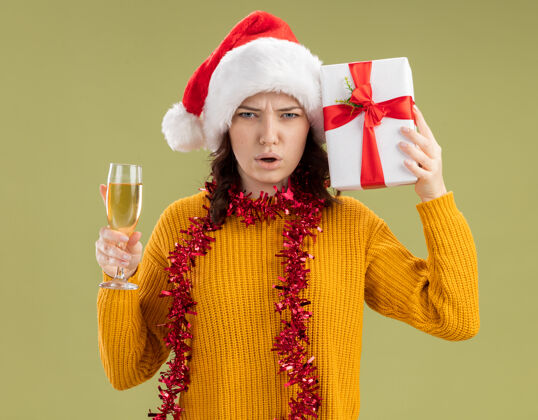 周围未出租的年轻斯拉夫女孩 戴着圣诞帽 脖子上戴着花环 手里拿着一杯香槟和圣诞礼盒 背景是橄榄绿 有复印空间帽子花环未出租