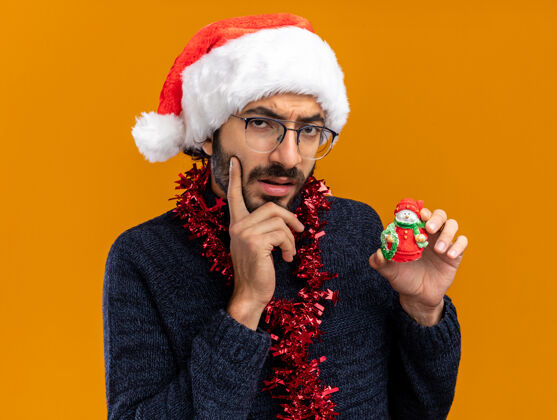 脸颊想着年轻帅哥戴着圣诞帽脖子上戴着花环拿着玩具把手指放在脸颊上孤立在橙色背景上思想手指抱