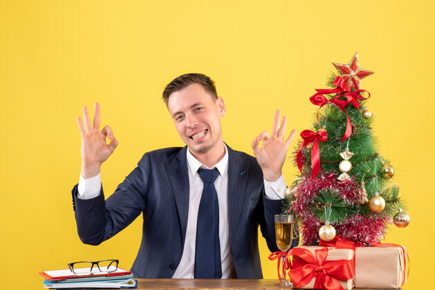 男人圣诞树旁的桌子旁 手拿着礼物摆在黄色的圣诞树上 快乐的男人正在做一个好的标志黄色商人行政人员