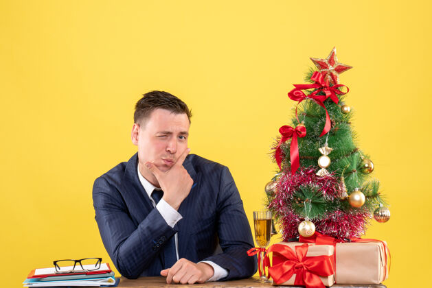 商人前视图中的男子穿着西装 眨着眼睛坐在圣诞树附近的桌子上 黄色的礼物公司眨眼男人