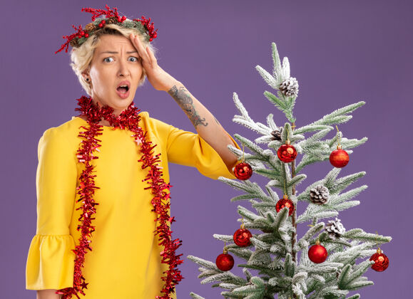 穿着戴着圣诞花环 脖子上戴着金属丝花环的金发女郎站在装饰好的圣诞树旁 手挽着手 孤零零地站在紫色的墙上保持花环关心