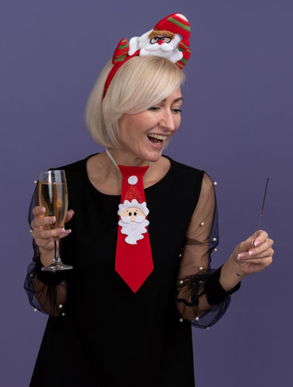 头带快乐的中年金发女人戴着圣诞老人的头带 打着领带 手里拿着节日的火花和一杯香槟 看着孤立在紫色墙上的火花中年穿着快乐