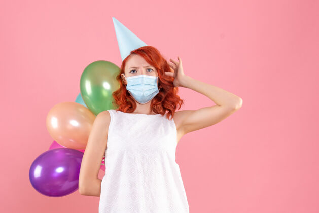 Covid前视图中的年轻女子拿着五颜六色的气球在无菌面具上粉红色的墙壁快乐快乐人