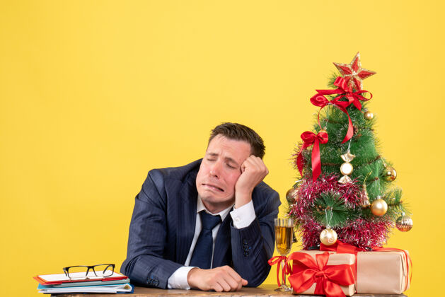 坐着悲伤的男人坐在圣诞树旁的桌子前 黄色的礼物西装职业男人