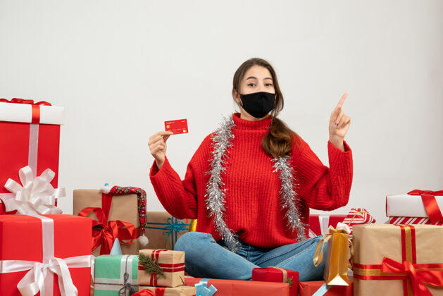 坐着一个穿着红色毛衣 戴着黑色面具的年轻女孩拿着卡片指着坐在旁边的东西 礼物是白色的年轻女孩面具红
