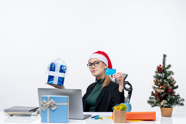 圣诞老人一个戴着圣诞老人帽子戴着眼镜的女商人坐在桌子旁 手里拿着圣诞礼物和银行卡礼物圣诞商务