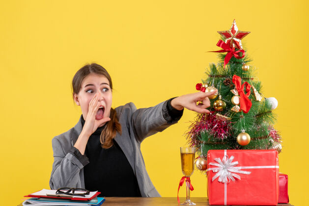 圣诞节前视图惊讶的女孩坐在办公桌旁大声喊叫 并在圣诞树和礼物鸡尾酒附近打电话给某人惊讶的女孩某人惊讶